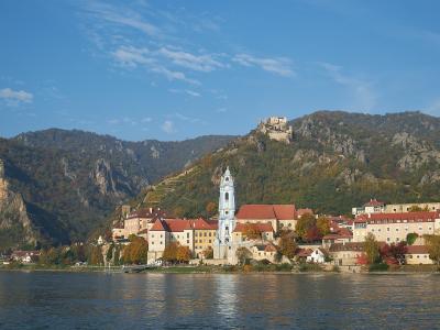 A scenic view of a town along a wide river featuring a prominent white church tower with a blue dome, terraced vineyards on the hillside, and a ruined castle on a rocky peak above.