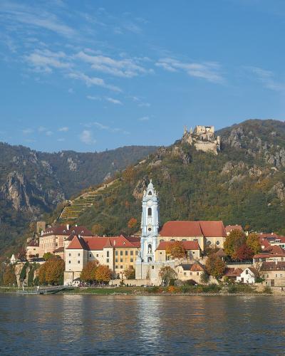 A scenic view of a town along a wide river featuring a prominent white church tower with a blue dome, terraced vineyards on the hillside, and a ruined castle on a rocky peak above.