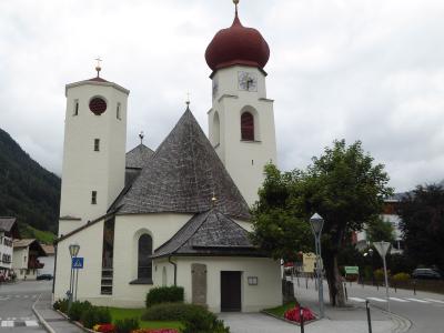 A white church with two towers (one square, one octagonal with a red onion dome) and a central shingled roof, situated at a street corner with mountains in the background.
