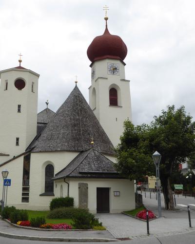 A white church with two towers (one square, one octagonal with a red onion dome) and a central shingled roof, situated at a street corner with mountains in the background.