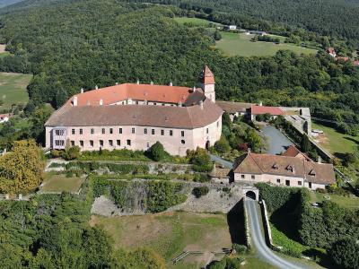 An aerial photograph of a large, historic castle complex with terracotta-tiled roofs, situated on a hilltop surrounded by dense green forest and agricultural fields, featuring a prominent tower, stone walls, and a swimming pool in the lower right.
