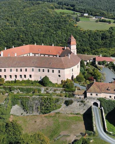 An aerial photograph of a large, historic castle complex with terracotta-tiled roofs, situated on a hilltop surrounded by dense green forest and agricultural fields, featuring a prominent tower, stone walls, and a swimming pool in the lower right.