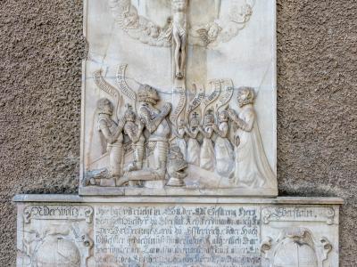 A close-up photograph of a stone memorial plaque mounted on a rough-textured wall, featuring a relief carving of the crucifixion above an inscription and two coats of arms.