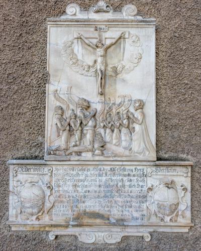 A close-up photograph of a stone memorial plaque mounted on a rough-textured wall, featuring a relief carving of the crucifixion above an inscription and two coats of arms.