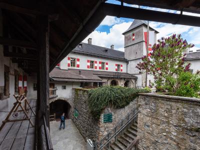 A view of a castle courtyard featuring white buildings with red-and-white striped shutters, a stone tower with a flag, and a person walking near an arched entrance.