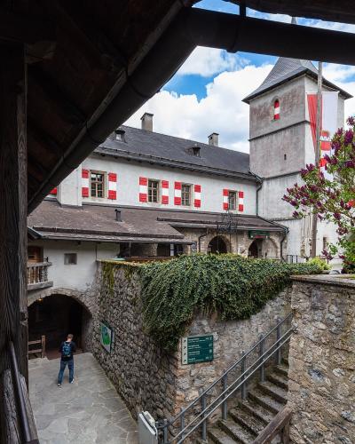 A view of a castle courtyard featuring white buildings with red-and-white striped shutters, a stone tower with a flag, and a person walking near an arched entrance.