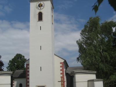 A tall, white church tower with a dark green spire, clock face, and red brick quoins, set against a blue sky with trees and a white gate in the foreground.