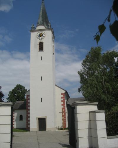 A tall, white church tower with a dark green spire, clock face, and red brick quoins, set against a blue sky with trees and a white gate in the foreground.