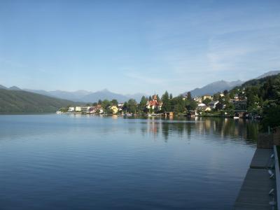 A scenic view of a lake with a town on the far shore featuring a prominent church with a red-tiled roof and a tower, set against a backdrop of mountains, with a wooden pier or walkway visible in the foreground right.