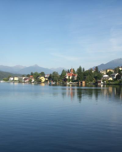 A scenic view of a lake with a town on the far shore featuring a prominent church with a red-tiled roof and a tower, set against a backdrop of mountains, with a wooden pier or walkway visible in the foreground right.