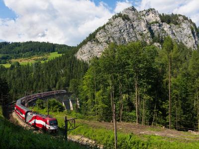 A large wooden picture frame stands in a forest, framing a view of a mountain valley containing viaducts and tunnels of a railway line.