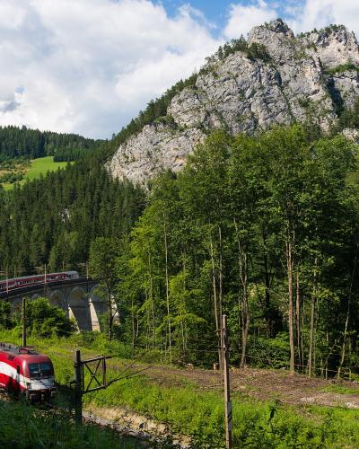 A large wooden picture frame stands in a forest, framing a view of a mountain valley containing viaducts and tunnels of a railway line.